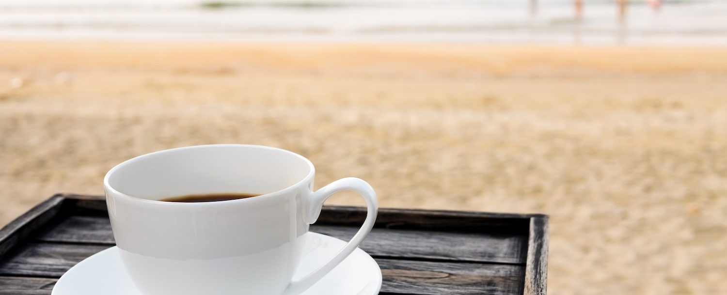 Close up white coffee cup on wood table at sunrise sand beach in the morning.