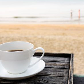 Close up white coffee cup on wood table at sunrise sand beach in the morning.
