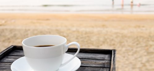 Close up white coffee cup on wood table at sunrise sand beach in the morning.