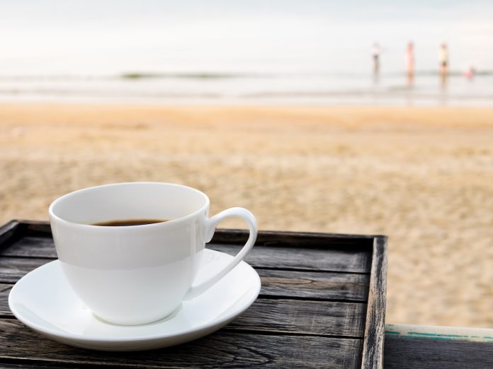 Close up white coffee cup on wood table at sunrise sand beach in the morning.