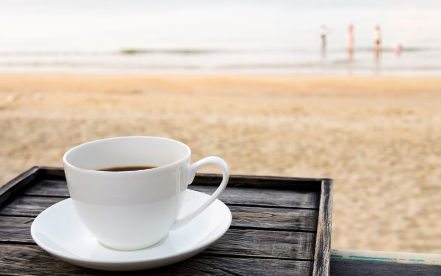 Close up white coffee cup on wood table at sunrise sand beach in the morning.
