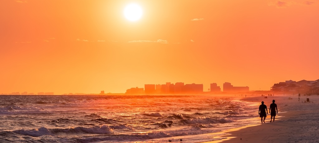 Couple walking on shore with dramatic orange red yellow sunset in Santa Rosa Beach, Florida with Pensacola coastline coast cityscape skyline in panhandle with ocean gulf mexico waves