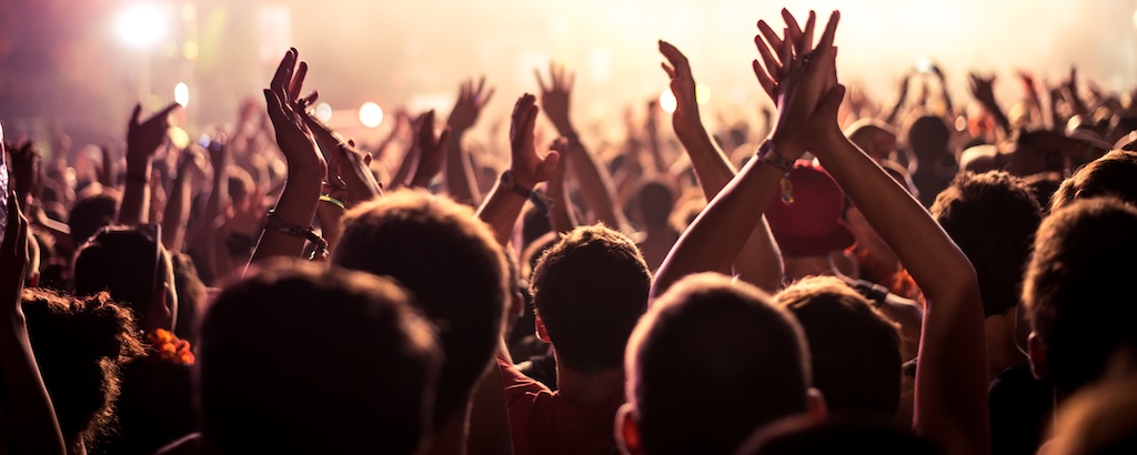 Audience with hands raised at a music festival and lights streaming down from above the stage. Soft focus, blurred movement.