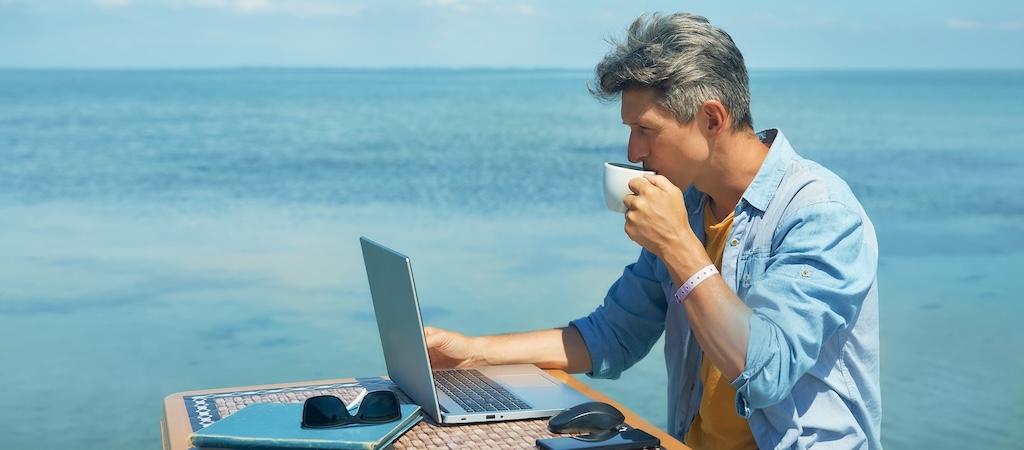 A freelancer working on a laptop computer at the beach by the blue sea, drinking coffee.