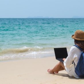 A traveler using and working on a laptop computer while sitting on the beach.
