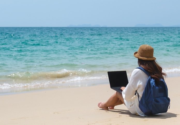 A traveler using and working on a laptop computer while sitting on the beach.