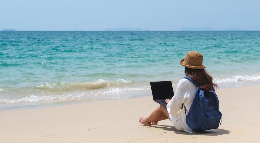 A traveler using and working on a laptop computer while sitting on the beach.