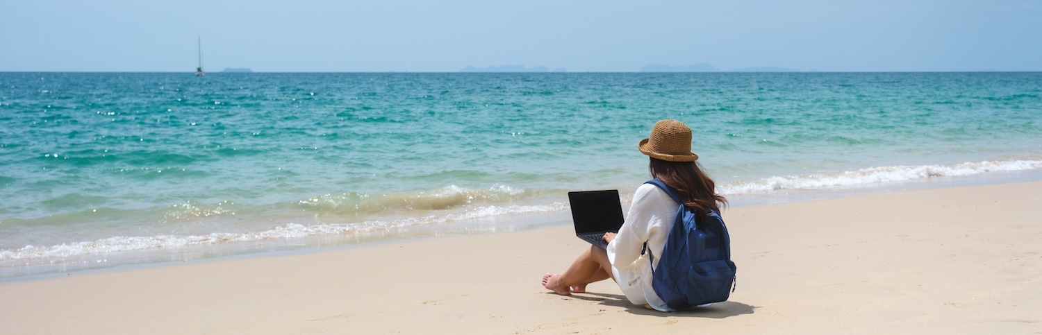 A traveler using and working on a laptop computer while sitting on the beach.